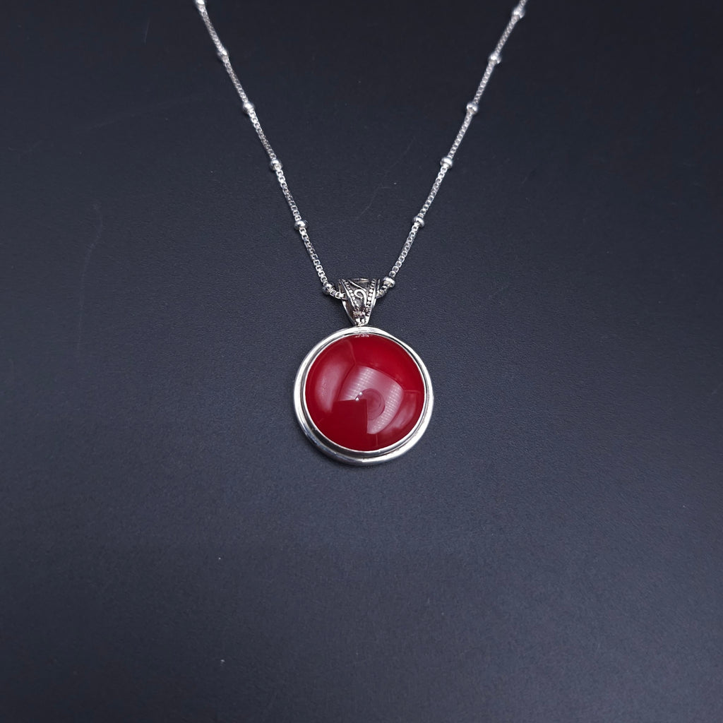 A silver necklace with a round orangish-red cabochon pendant and a fancy swirly oxidized bail. On a silver station chain with a silver bead every 1/2"