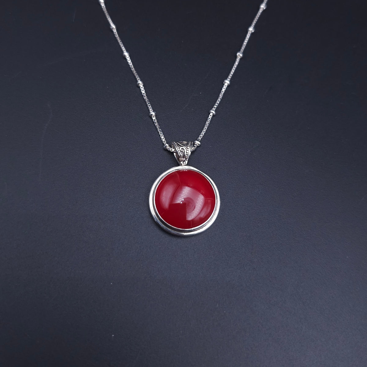 A silver necklace with a round orangish-red cabochon pendant and a fancy swirly oxidized bail. On a silver station chain with a silver bead every 1/2"