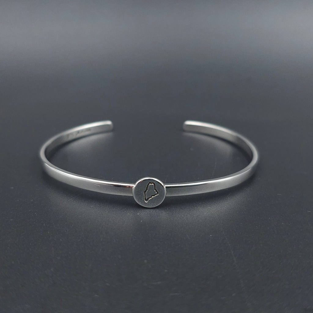 A polished silver cuff bracelet with a small circular charm featuring an engraved outline of the state of Maine attached to the center of the cuff.