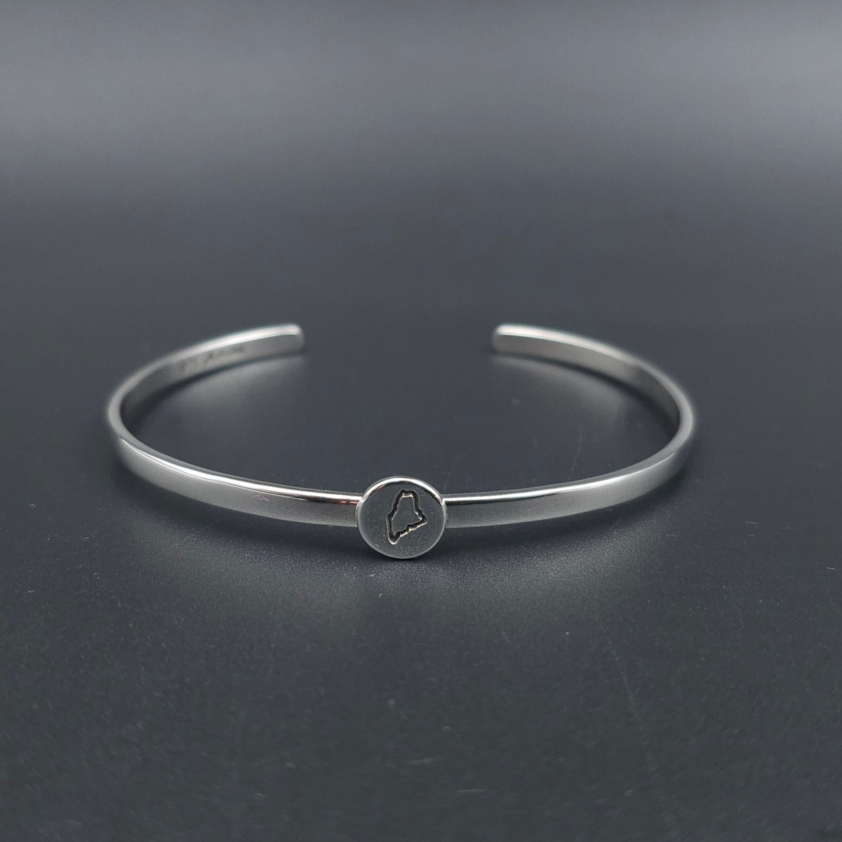 A polished silver cuff bracelet with a small circular charm featuring an engraved outline of the state of Maine attached to the center of the cuff.