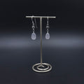 Silver dangle earrings with flower charms hanging from French earrings wires, and then light purple sea glass hanging from the flower charms.
