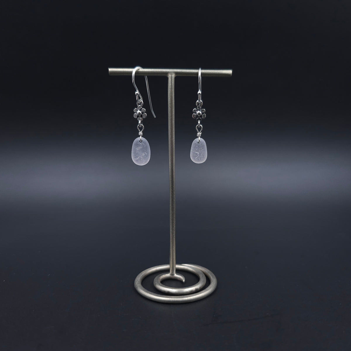 Silver dangle earrings with flower charms hanging from French earrings wires, and then light purple sea glass hanging from the flower charms.
