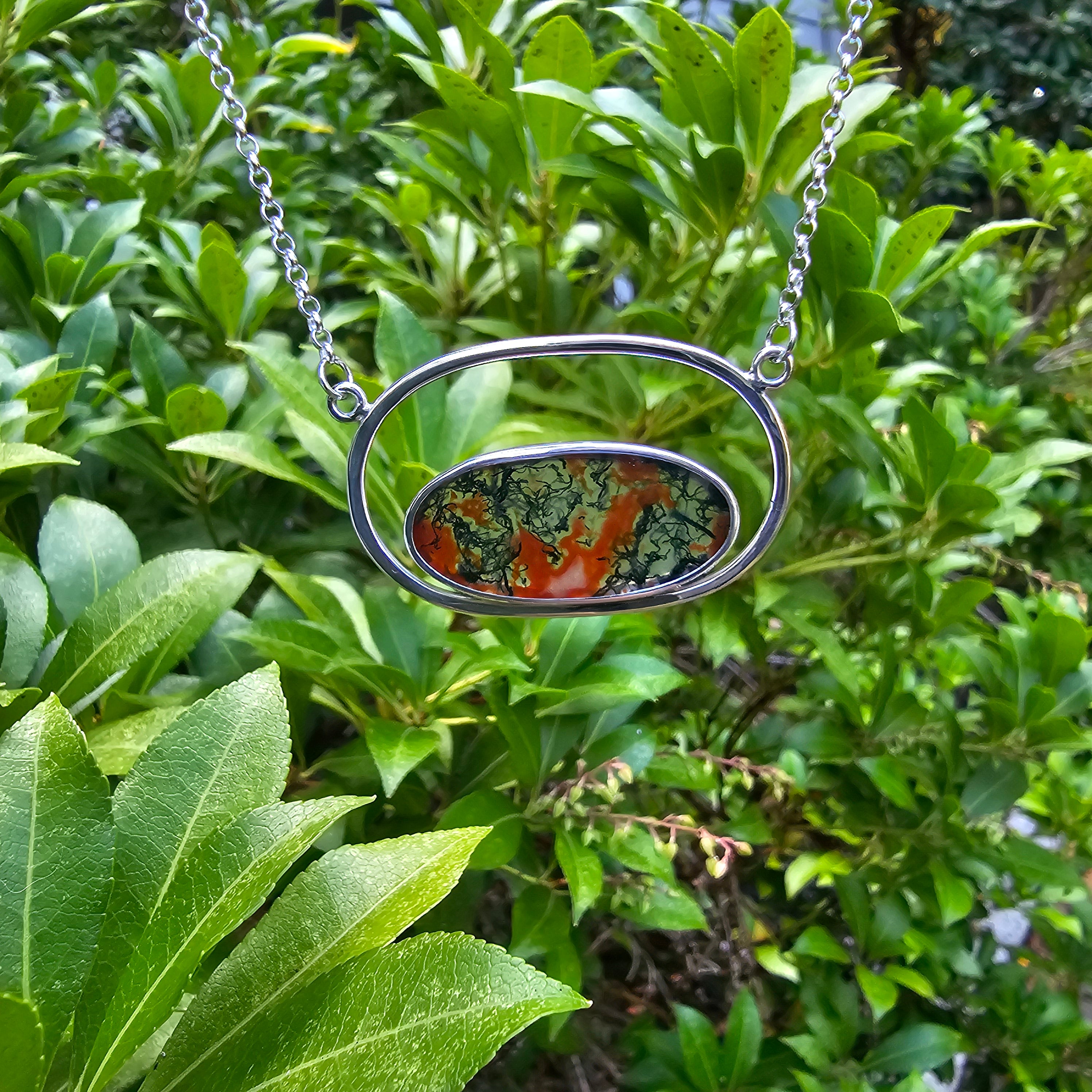 horizontal oval silver wire pendant with bezel set long oval moss agate stone inside. Stone has green and orange inclusions.