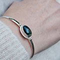 A sterling silver cuff bracelet with a dark green oval moss agate cabochon stone with a textured silver border and a silver ball on each side of stone.
