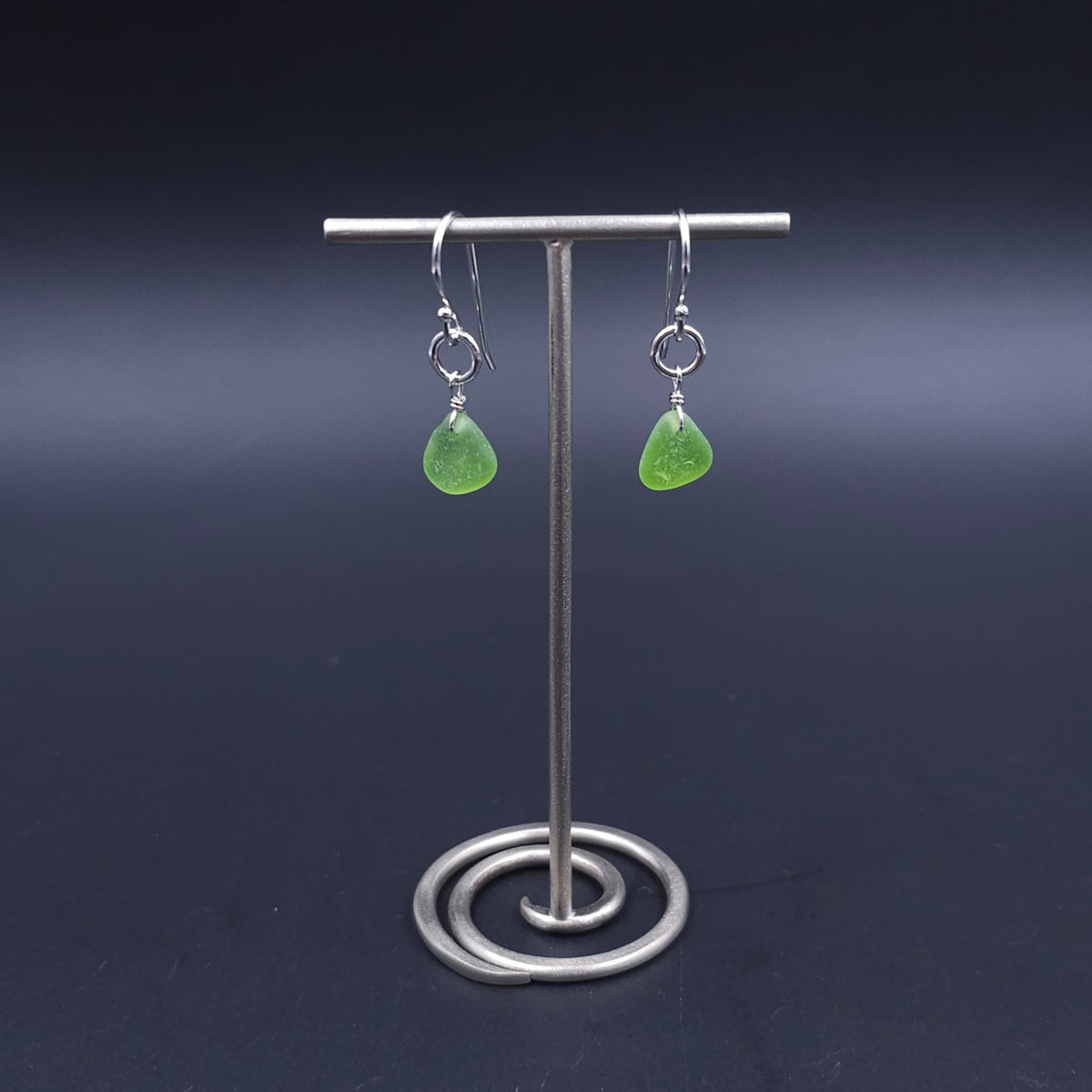 A pair of silver dangle earrings with small green teardrop-shaped sea glass, hanging from small silver circles.