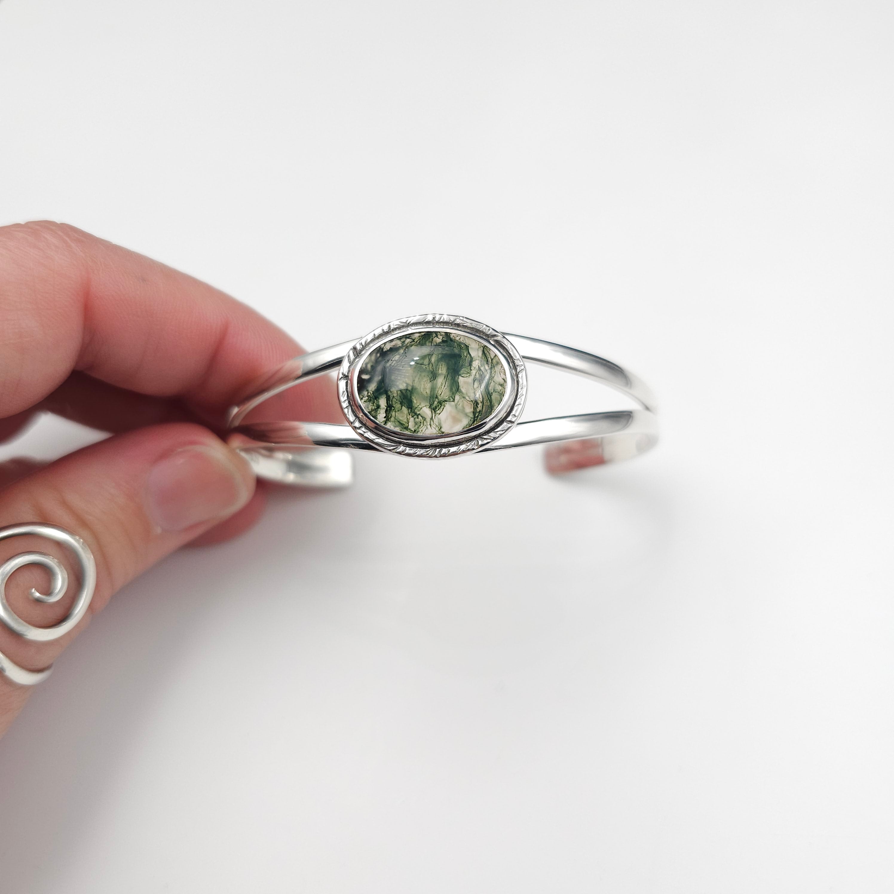 Sterling cuff made from two pieces of half round wire connected in the back and open on the top. Bezel set stone with dark green and clear natural patterns. A textured silver border surrounds the stone.