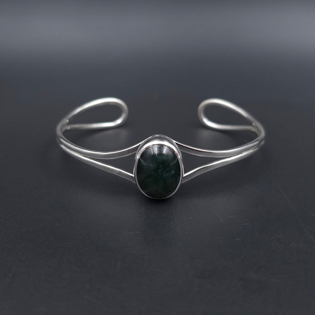 A silver-toned open cuff bracelet featuring a central oval bezel-set dark green stone, displayed on a dark gray background.