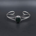 A silver-toned open cuff bracelet featuring a central oval bezel-set dark green stone, displayed on a dark gray background.