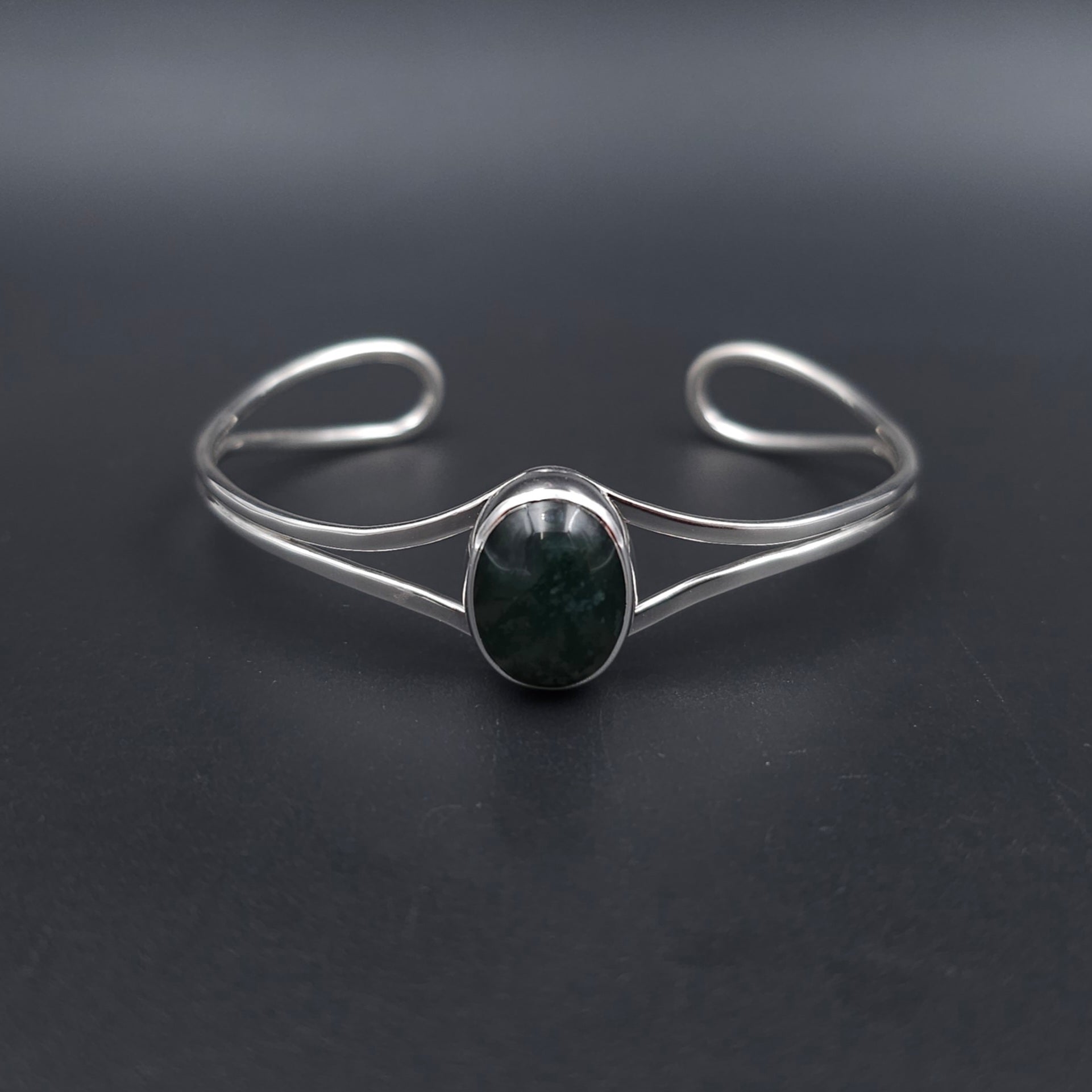 A silver-toned open cuff bracelet featuring a central oval bezel-set dark green stone, displayed on a dark gray background.
