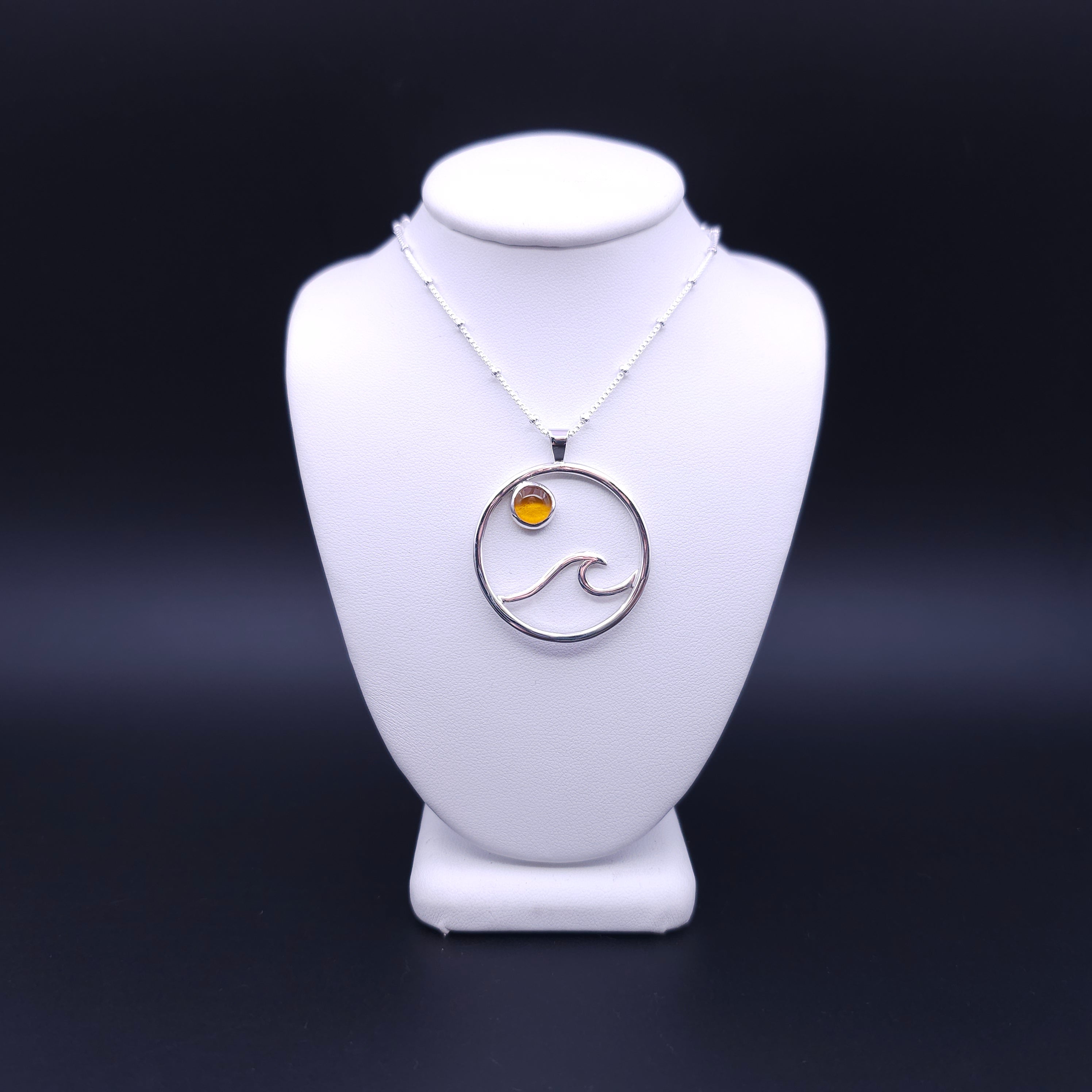 Silver circle pendant with wave made from wire and a round orange amber stone at top of circle slightly off center