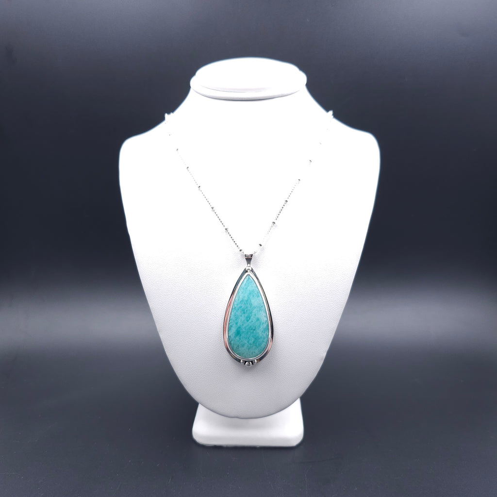 A silver teardrop pendant necklace with a turquoise-blue amazonite stone. Above the stone is one silver ball, and below are three silver balls, with the largest in the center. There is a slight silver sheet border around the whole stone. The chain is a silver box station chain with a silver bead every half inch.