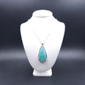 A silver teardrop pendant necklace with a turquoise-blue amazonite stone. Above the stone is one silver ball, and below are three silver balls, with the largest in the center. There is a slight silver sheet border around the whole stone. The chain is a silver box station chain with a silver bead every half inch.