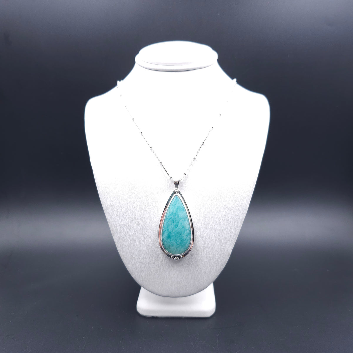 A silver teardrop pendant necklace with a turquoise-blue amazonite stone. Above the stone is one silver ball, and below are three silver balls, with the largest in the center. There is a slight silver sheet border around the whole stone. The chain is a silver box station chain with a silver bead every half inch.