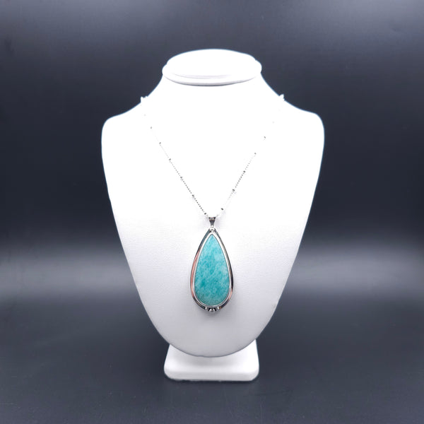 A silver teardrop pendant necklace with a turquoise-blue amazonite stone. Above the stone is one silver ball, and below are three silver balls, with the largest in the center. There is a slight silver sheet border around the whole stone. The chain is a silver box station chain with a silver bead every half inch.