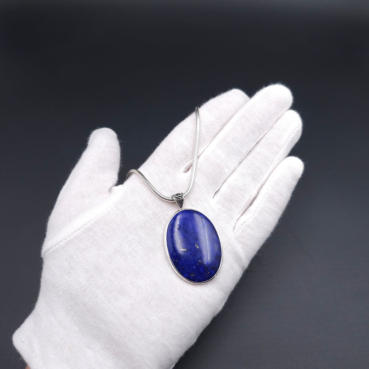 An oval silver pendant necklace with a blue lapis lazuli stone and an oxidized fancy bail, on a snake chain
