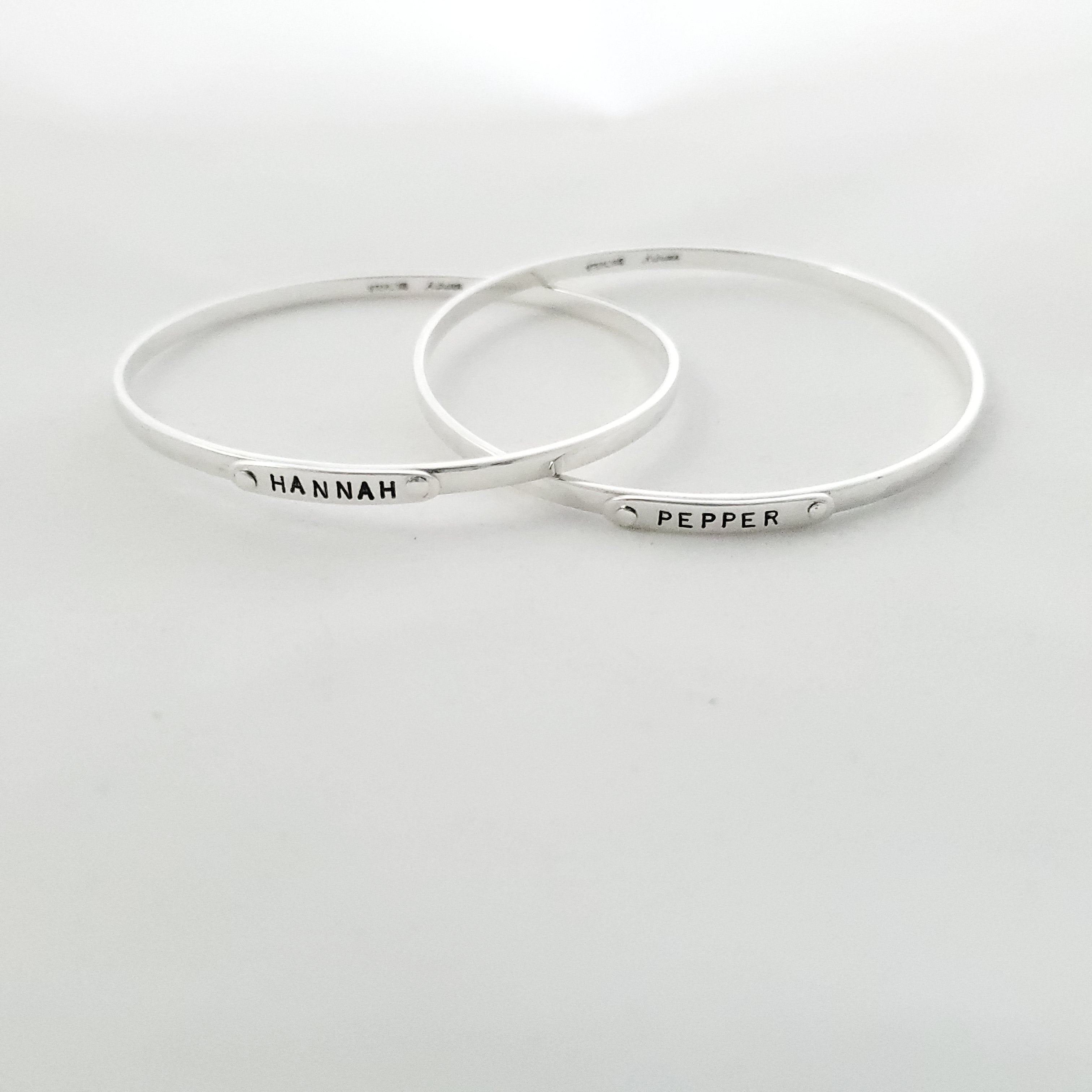 Two silver bangle bracelets shown spread apart but connected. One says HANNAH, the other says PEPPER.